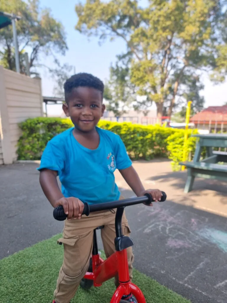 A confident Scamps preschooler riding a bicycle with training wheels outdoors